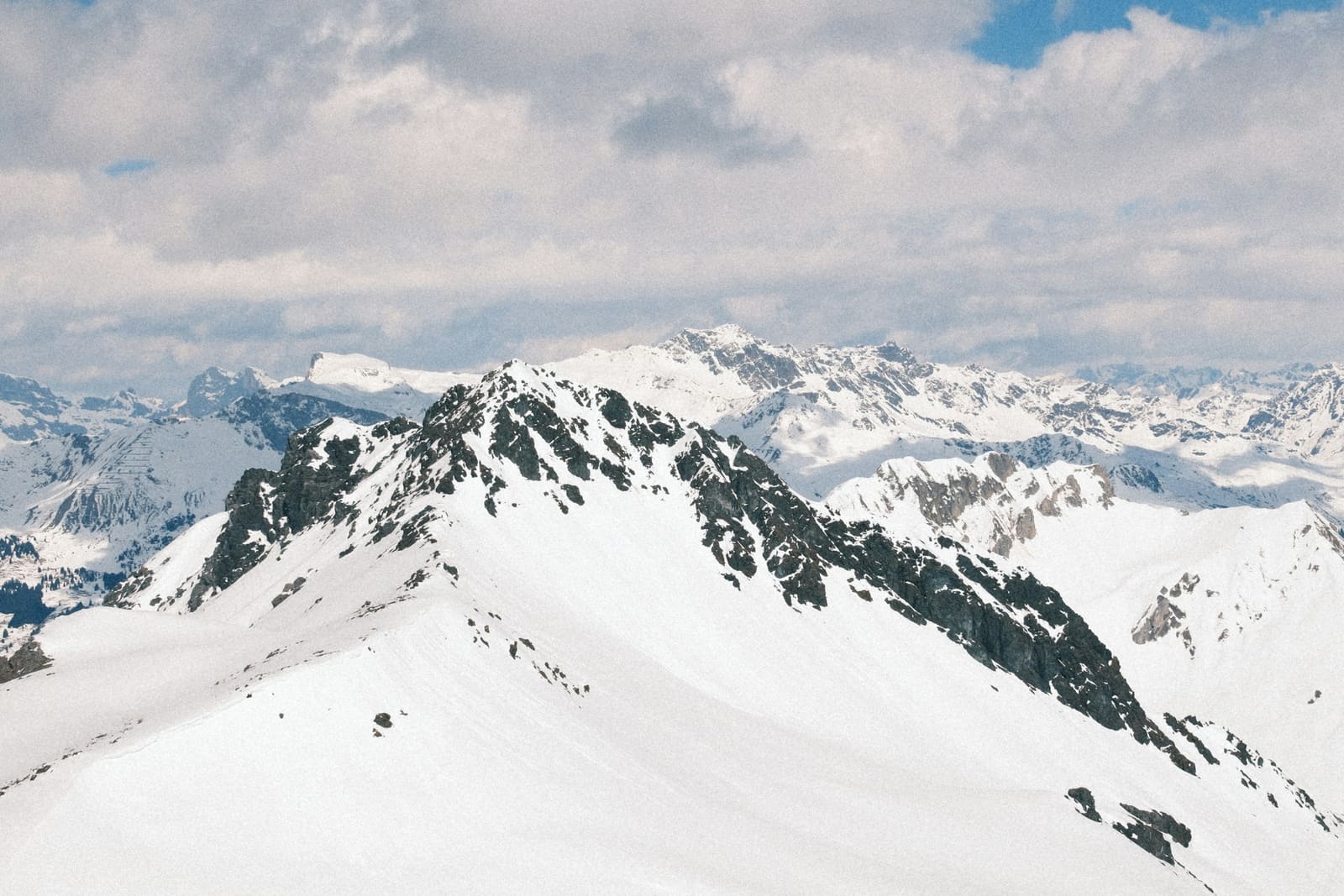 Verschneiter Bergkamm mit Felsen unter bewölktem Himmel und fernen Gipfeln