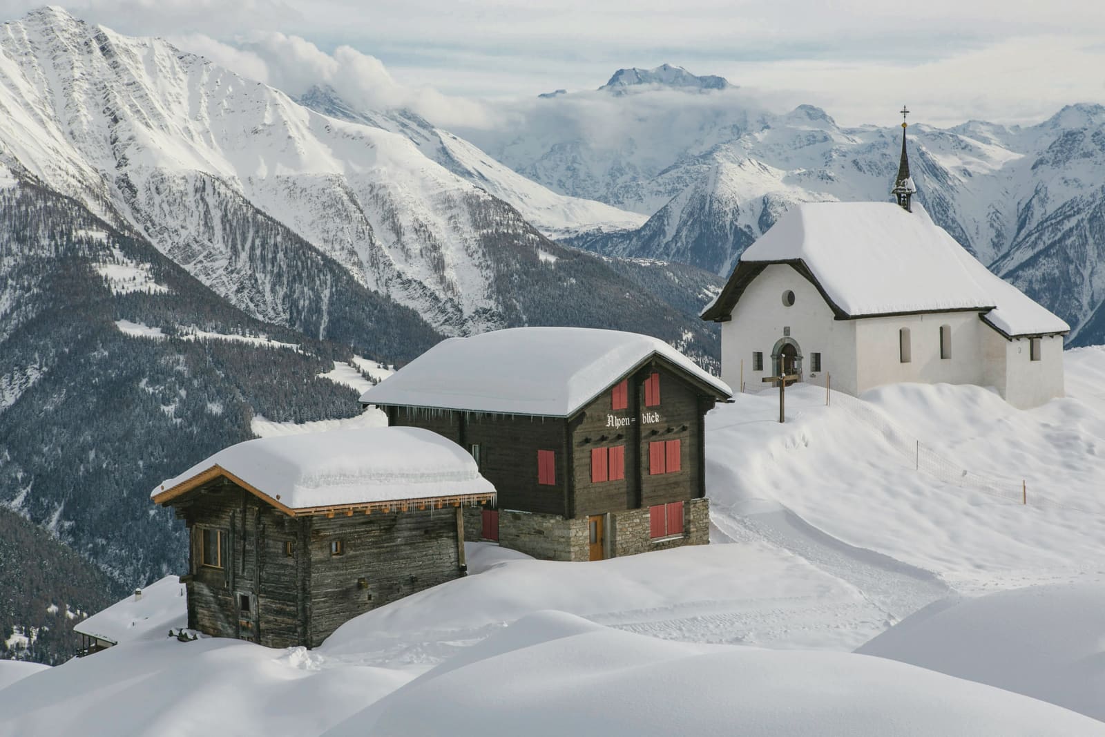 Verschneite Holzhäuser und eine weisse Kapelle hoch über dem Tal mit Bergpanorama