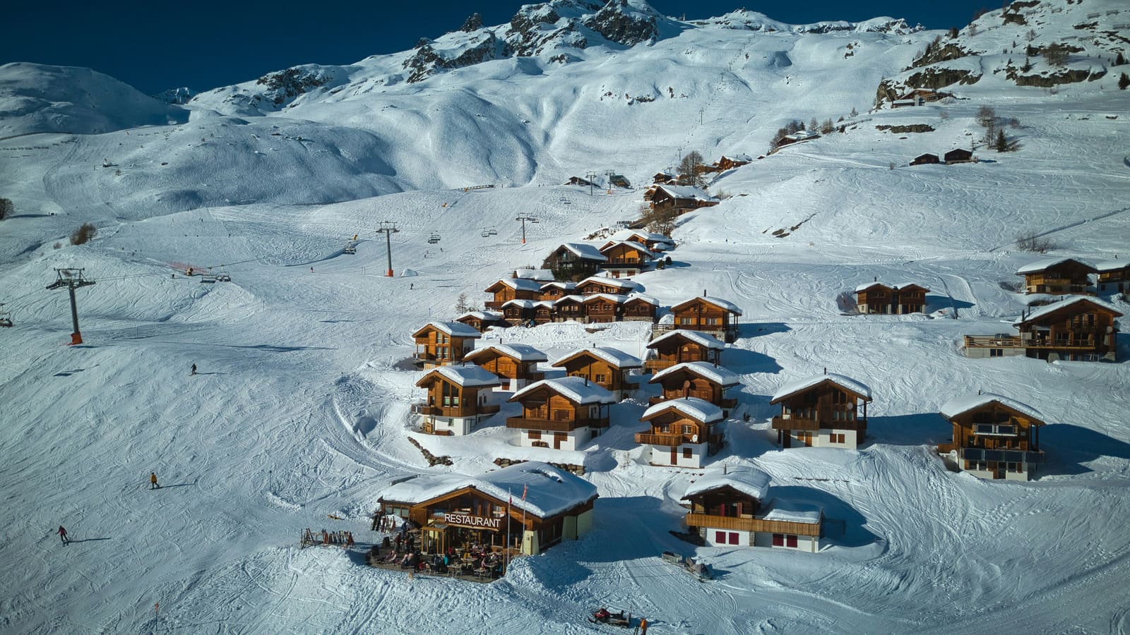 Verschneite Chaletsiedlung mit Bergrestaurant, Pisten und Liften aus der Luft