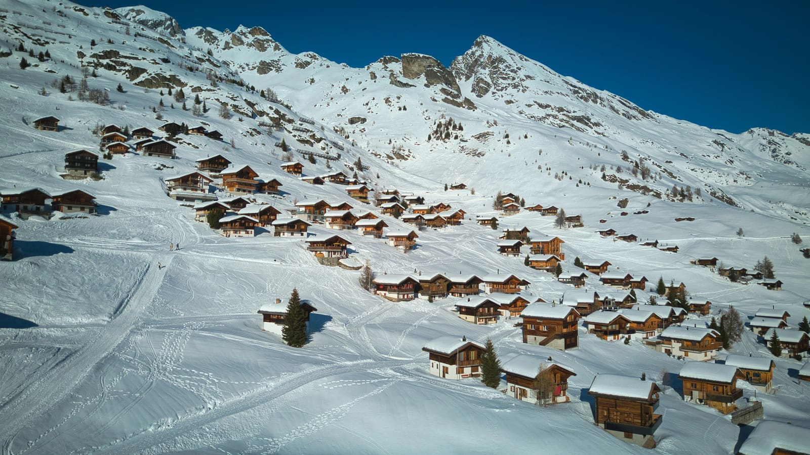 Dunkle Holzchalets verstreut an verschneiten Sonnenhängen unter tiefblauem Himmel