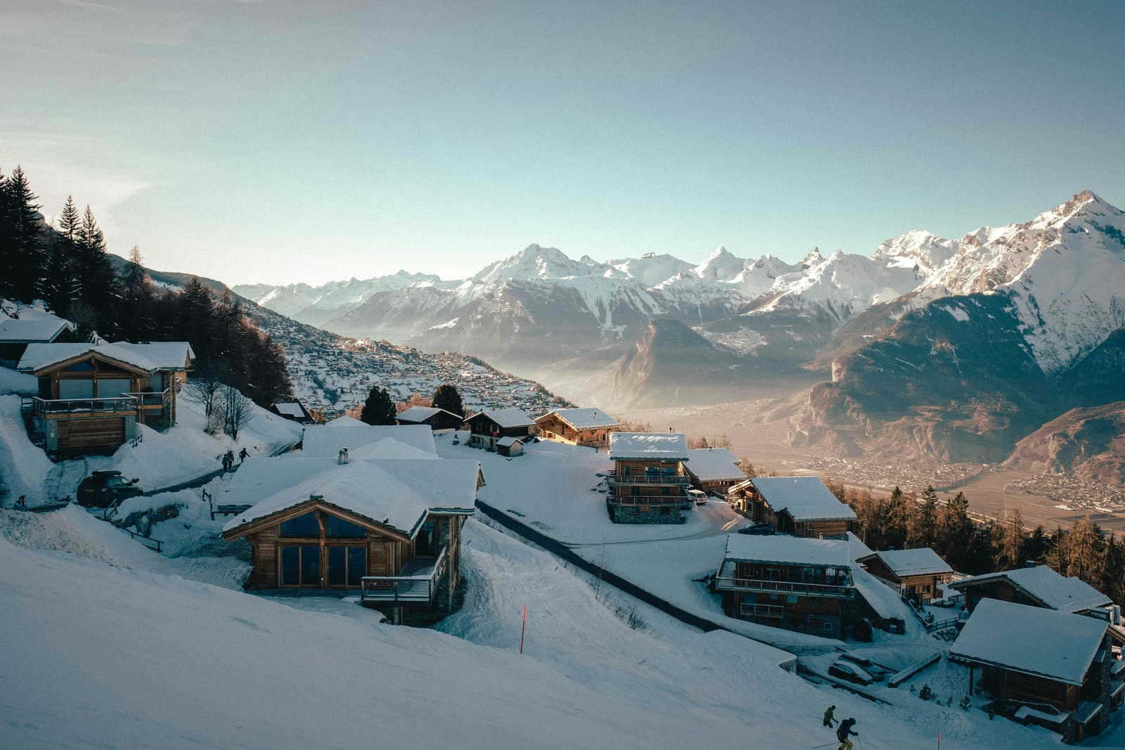 Holzchalets an einem verschneiten Hang im Morgenlicht über einem weiten Alpental