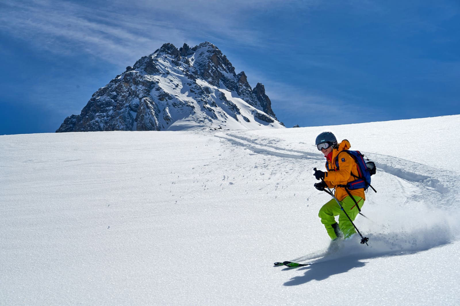 Freerider in oranger Jacke fährt durch Pulverschnee unter einem felsigen Gipfel
