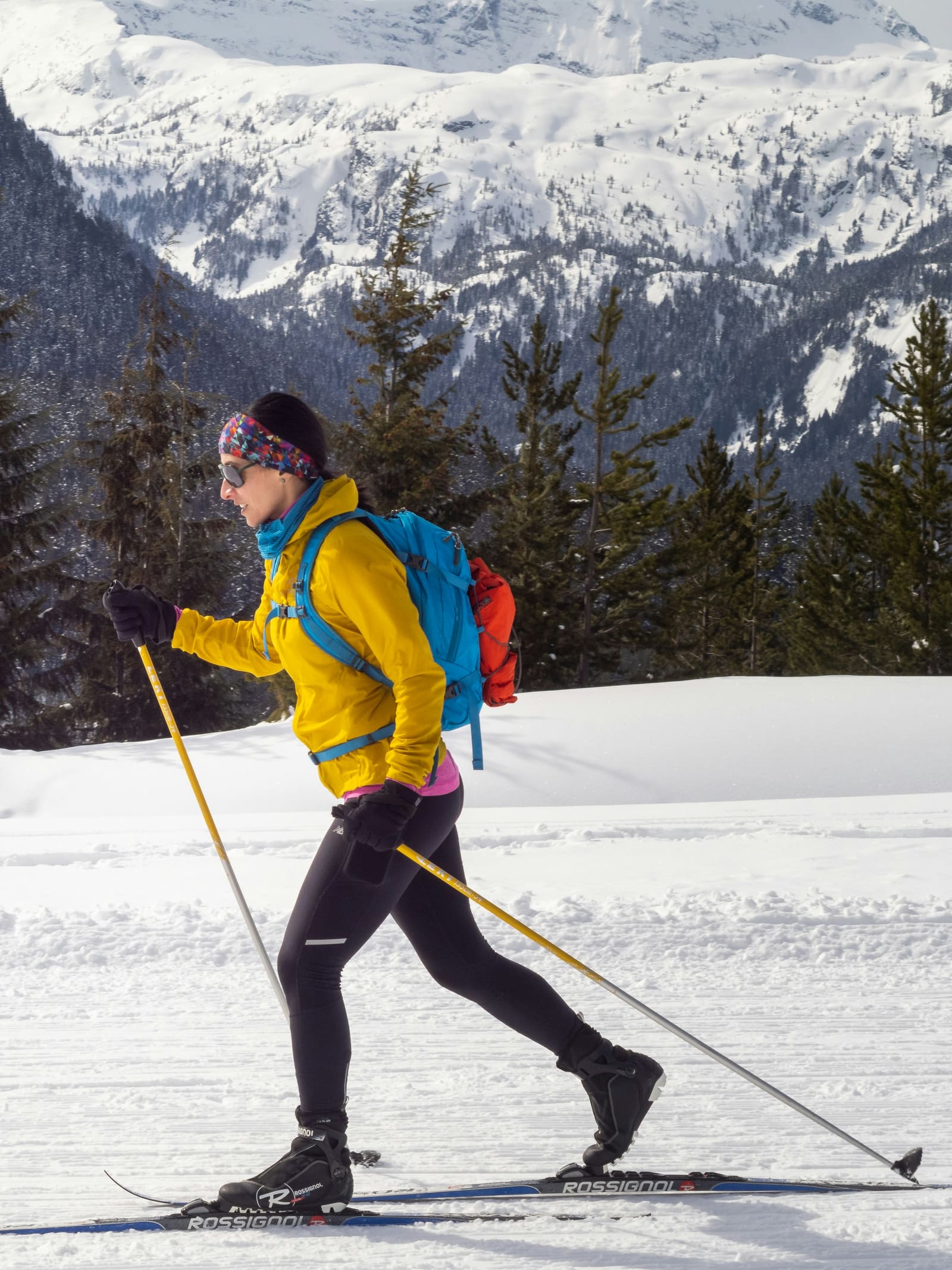 Eine Skitourengängerin in gelber Jacke mit Rucksack im Aufstieg vor verschneitem Wald.