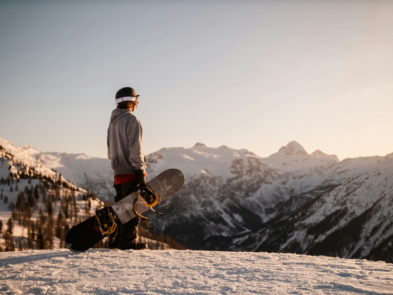 Snowboarder hält sein Brett und blickt im Abendlicht auf verschneite Berge