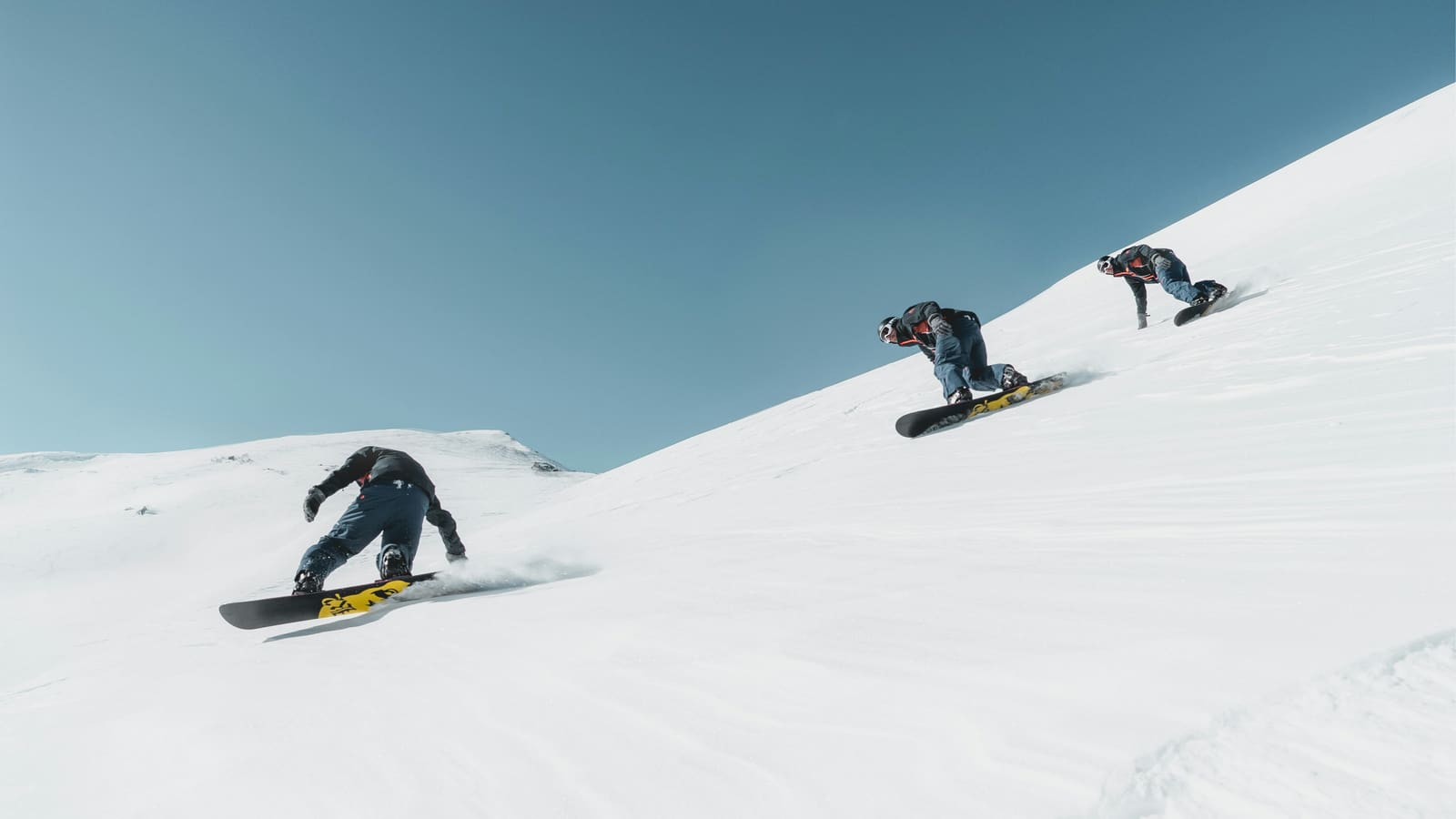 Drei Snowboarder carven hintereinander über einen weiten Hang unter blauem Himmel